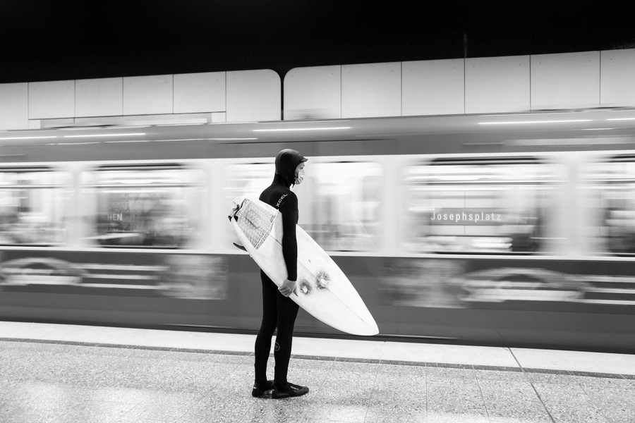 A man wearing a wetsuit and mask carries a surfboard under his arm, waiting as a train passes in a subway station.