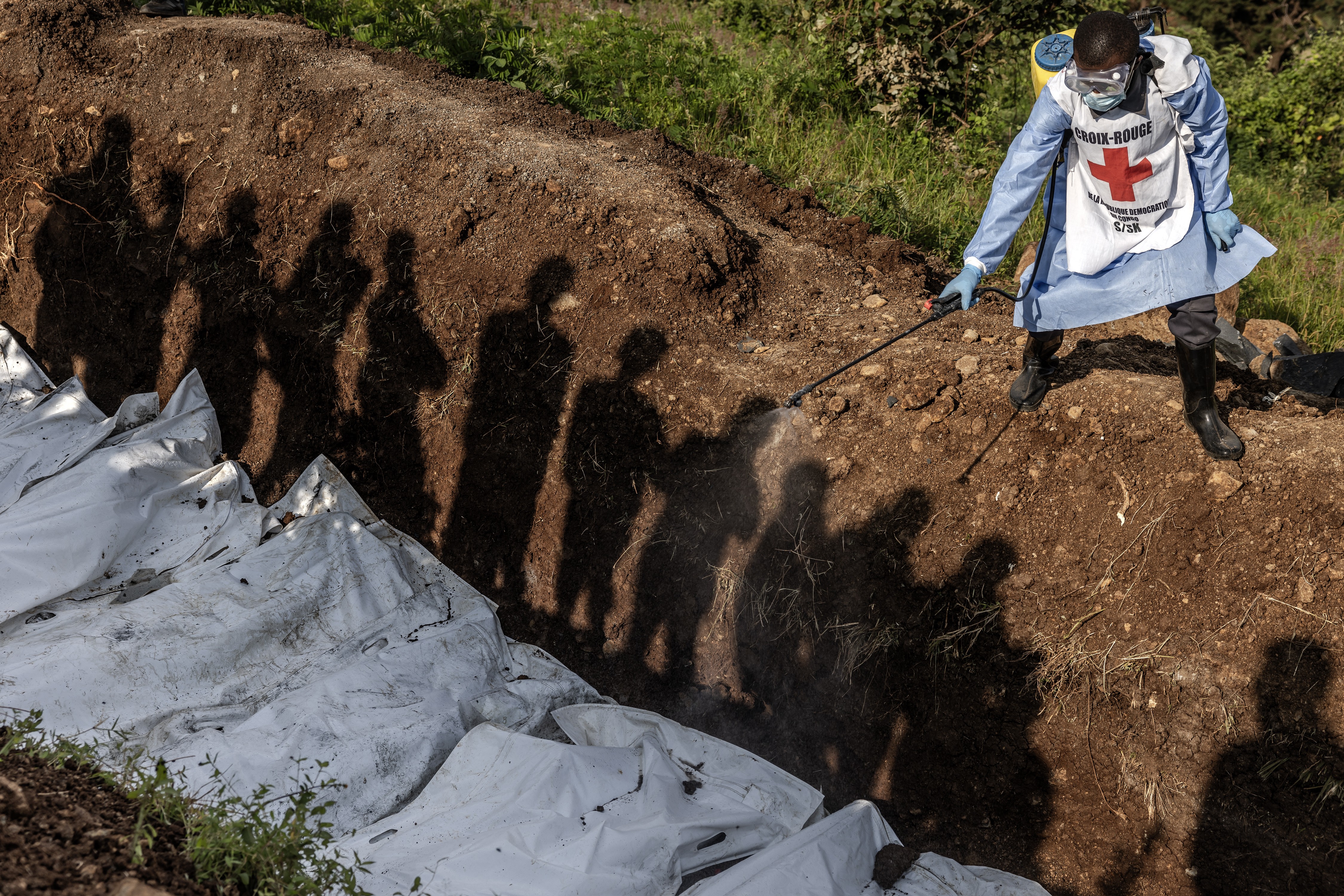 People's shadows fall along the wall of a mass grave, as a Red Cross worker sprays disinfectant on bodies in white bags.