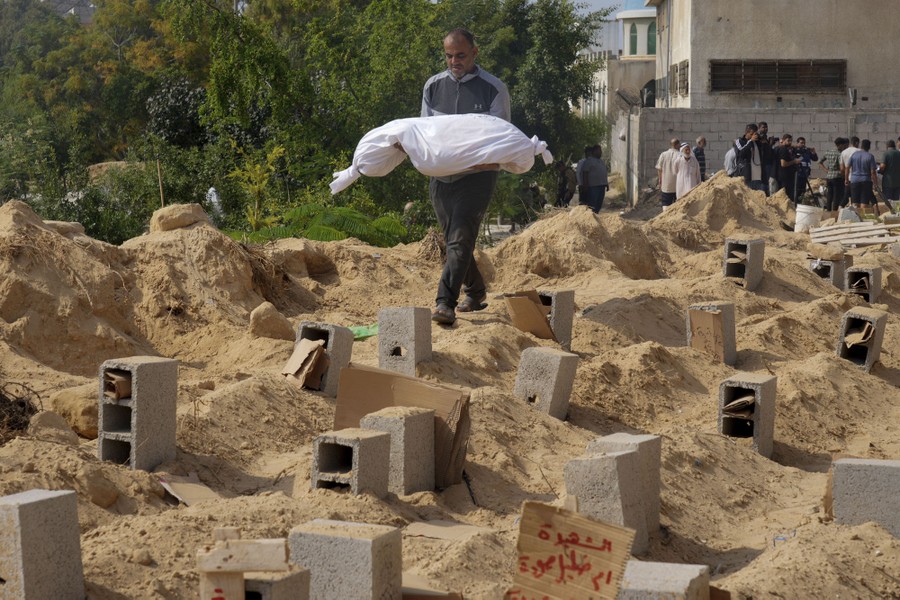 A person carries a small body, wrapped in a sheet, into a part of a cemetery with many newly dug graves.