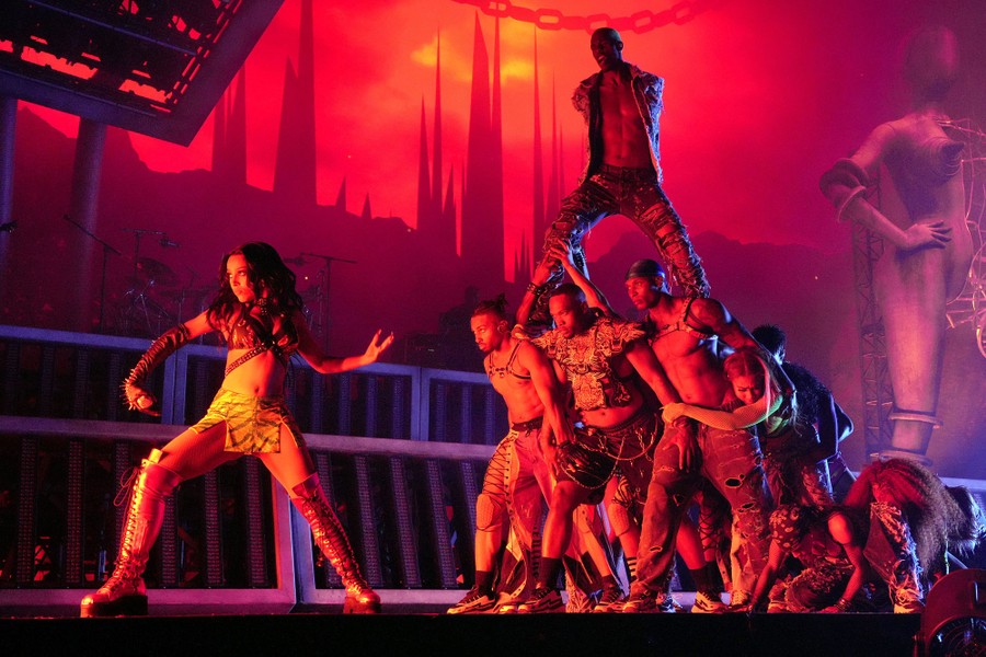 Performer Doja Cat strikes a pose while gesturing to about a dozen backup dancers standing together behind her.