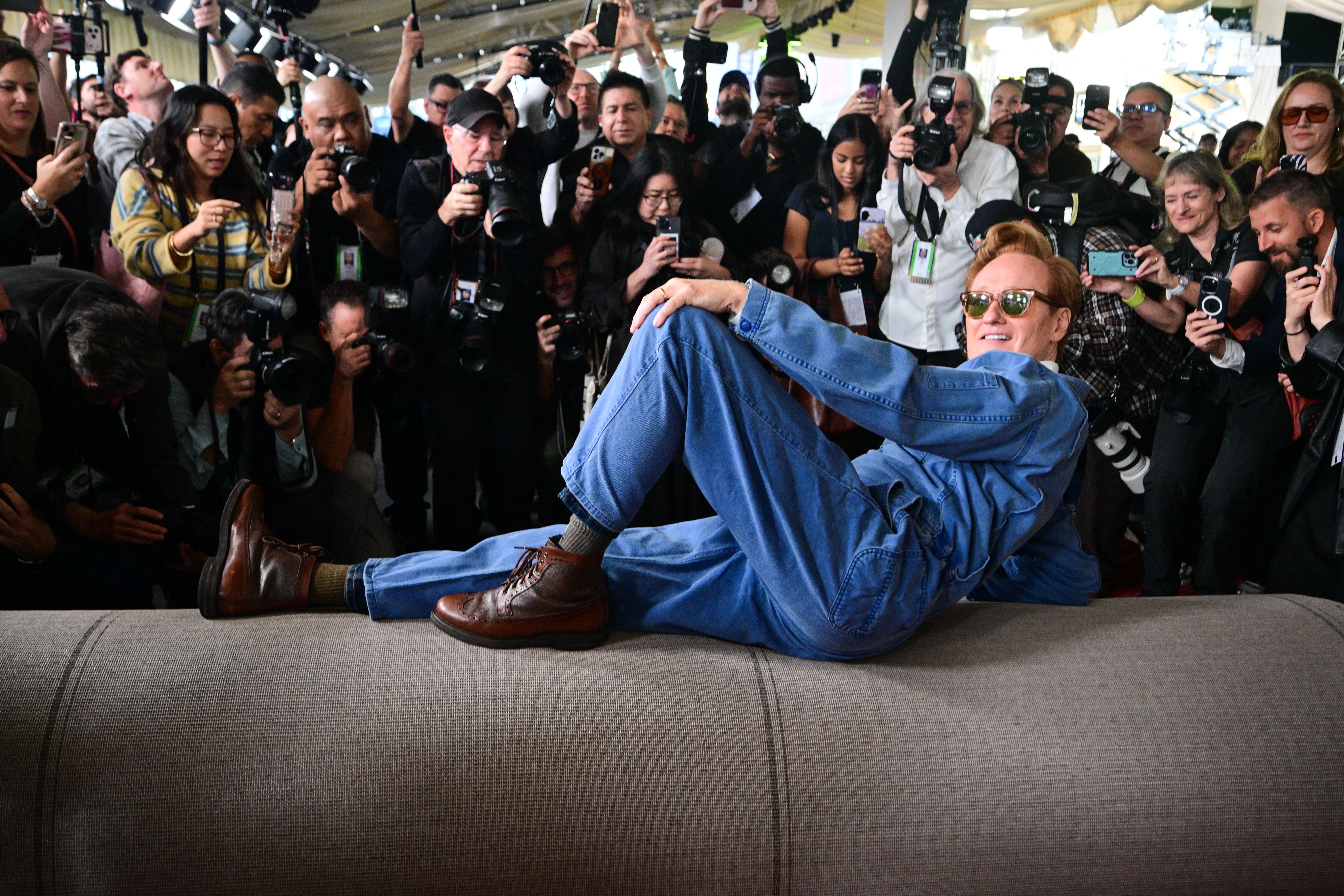The performer Conan O'Brien poses for a large group of photographers, lying on a rolled-up red carpet.