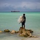 Kaitara Kautu, a net fisherman whose home flooded during last year's king tide in Betio, a town on the island of South Tarawa in Kiribati, stands on a rock in the middle of turquoise-colored water; a net rests on his left shoulder, and large ships sit in the distance.