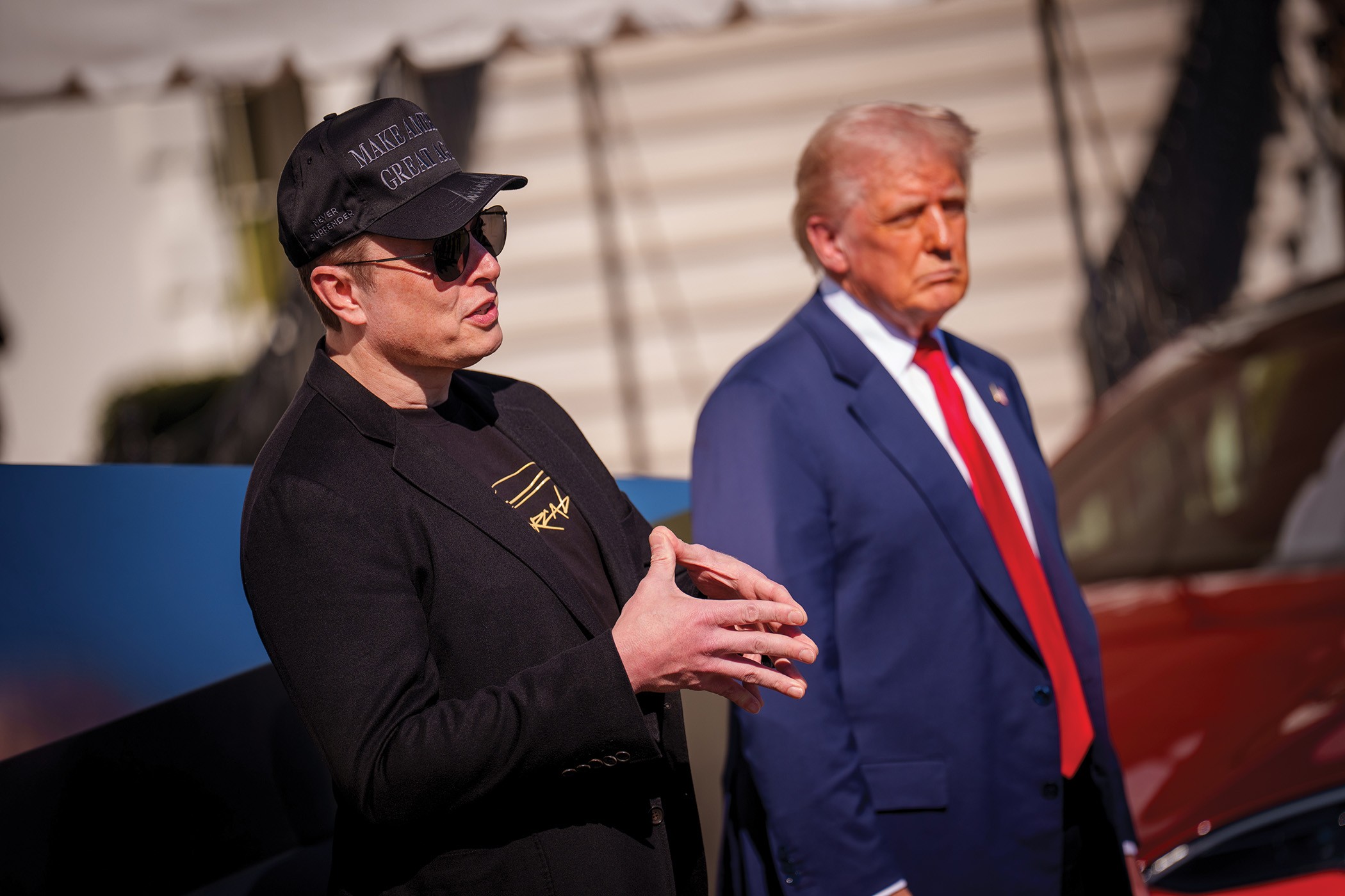 photo of Elon Musk wearing all-black 'Make America Great Again' baseball cap and sunglasses speaking at event next to Donald Trump in suit