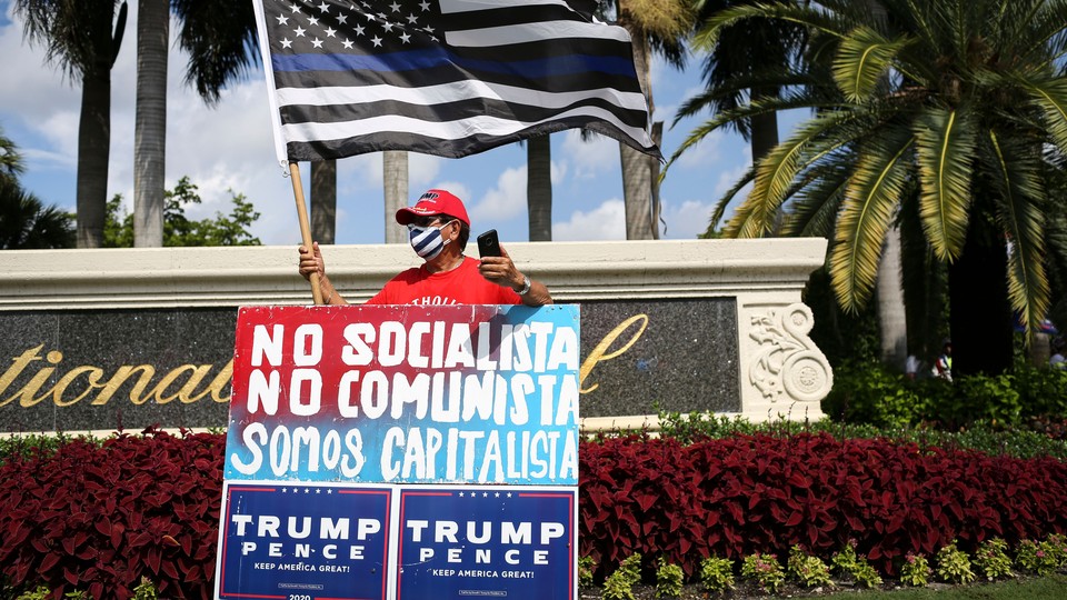 A man in a MAGA hat stands with a sign that reads "Not socialist, not communist, we are capitalist" in Spanish