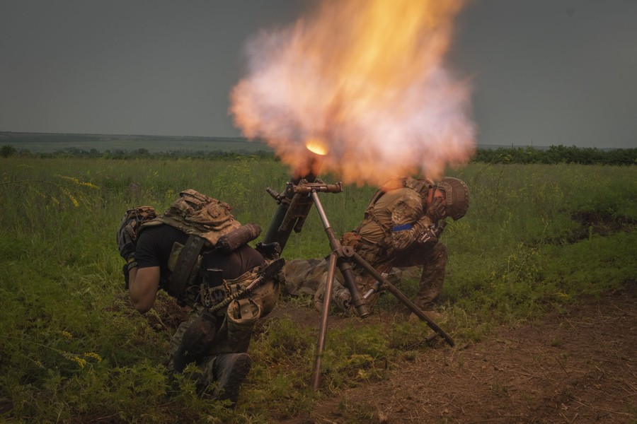 Two soldiers kneel and cover their ears as a mortar launch takes place, creating a small fireball.