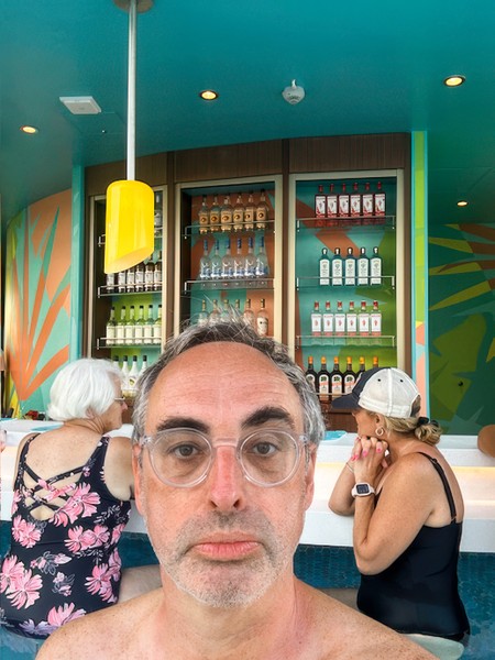 selfie photo of man with glasses, in background is swim-up bar with two women facing away