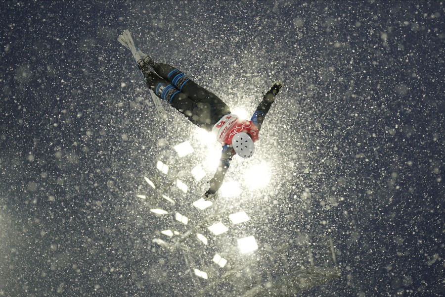 A skier is seen in mid-air during a jump, backlit by spotlights, amid a snowstorm.
