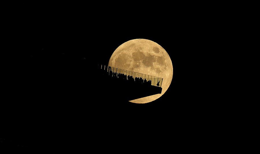 Silhouettes of people standing on a high observation deck in front of the full moon