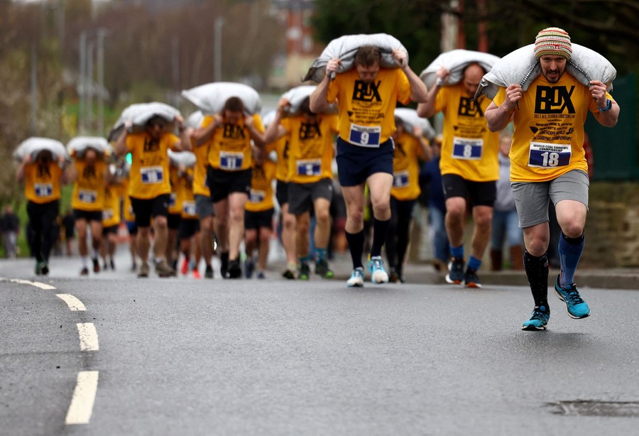 Dozens of men wearing shorts and yellow T-shirts run in a race while carrying heavy bags of coal over their shoulders.