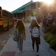 Two girls walk to school. A school bus is on their left, and a group of children wearing masks is on the right.