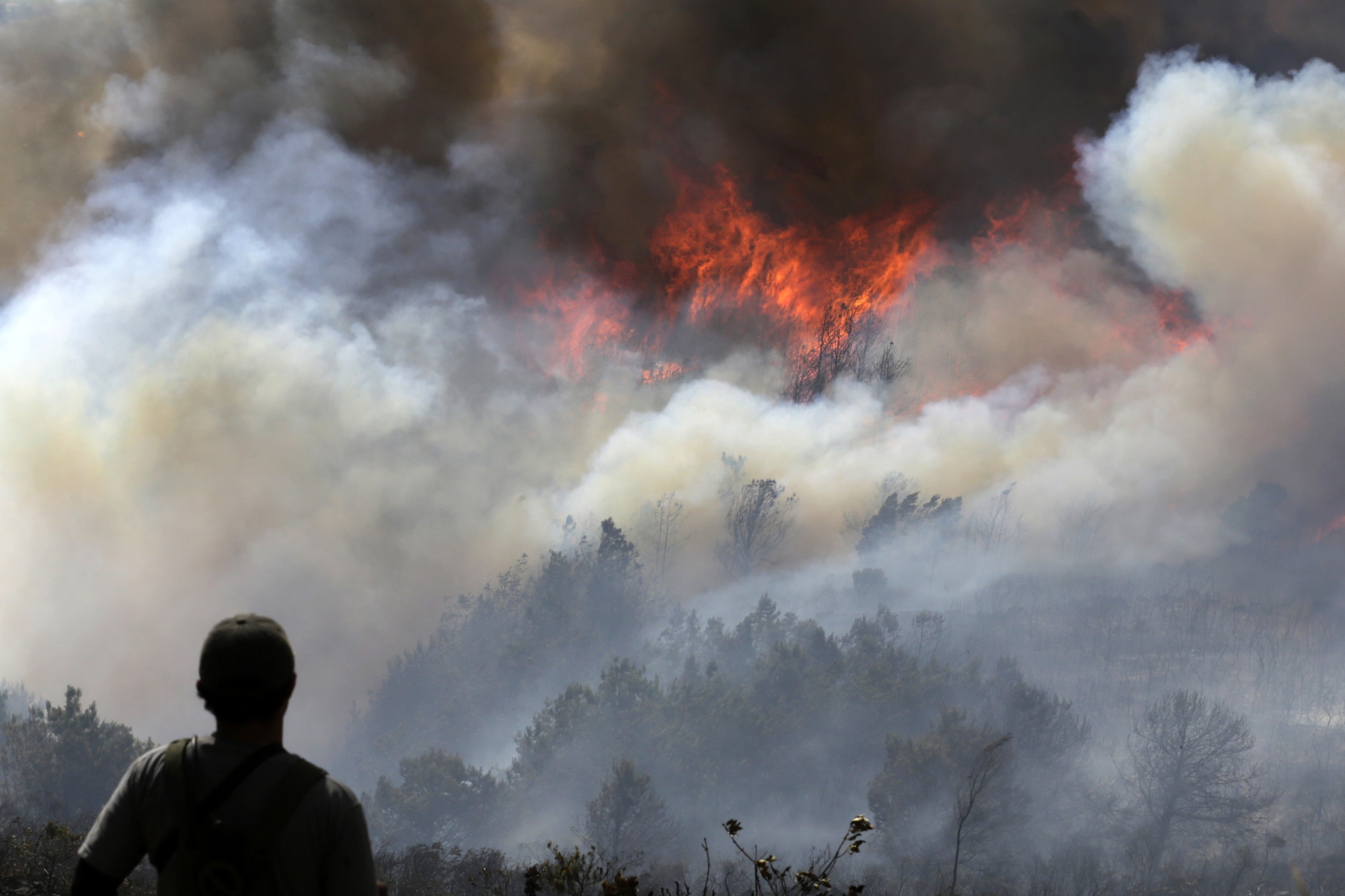 A person looks up toward a raging forest fire on a hillside, partially obscured by thick smoke.