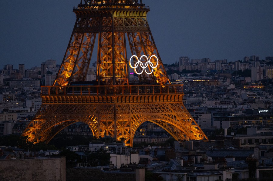 An elevated view of Paris buildings and part of the Eiffel Tower, with the Olympic rings mounted on the tower's side