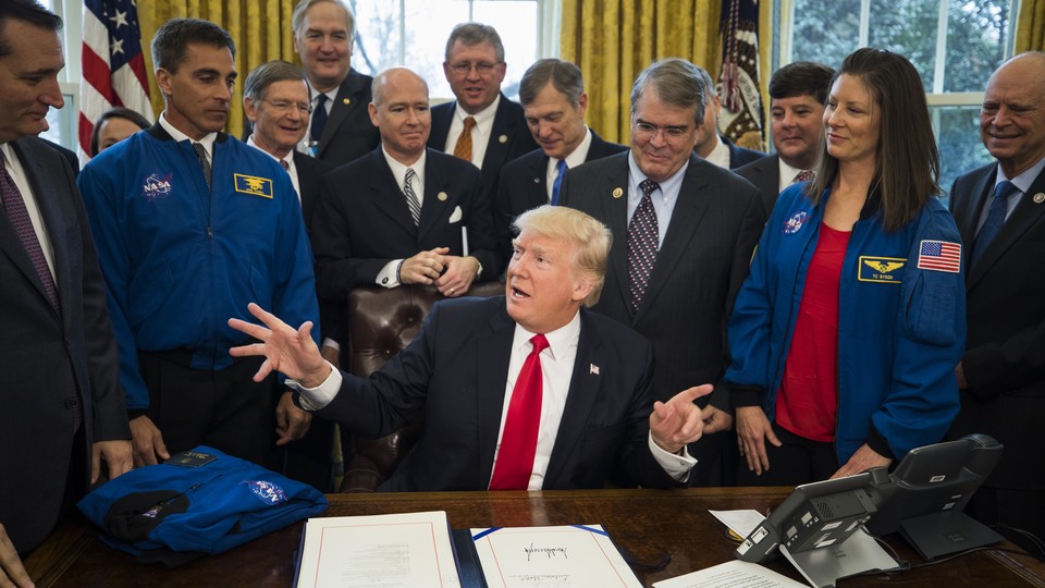 President Trump sits at the Resolute Desk surrounded by men in suits and two people in NASA jackets.