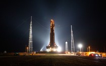 A night view of a tall, illuminated rocket and its support structure on a mobile platform, standing amid several tall towers.