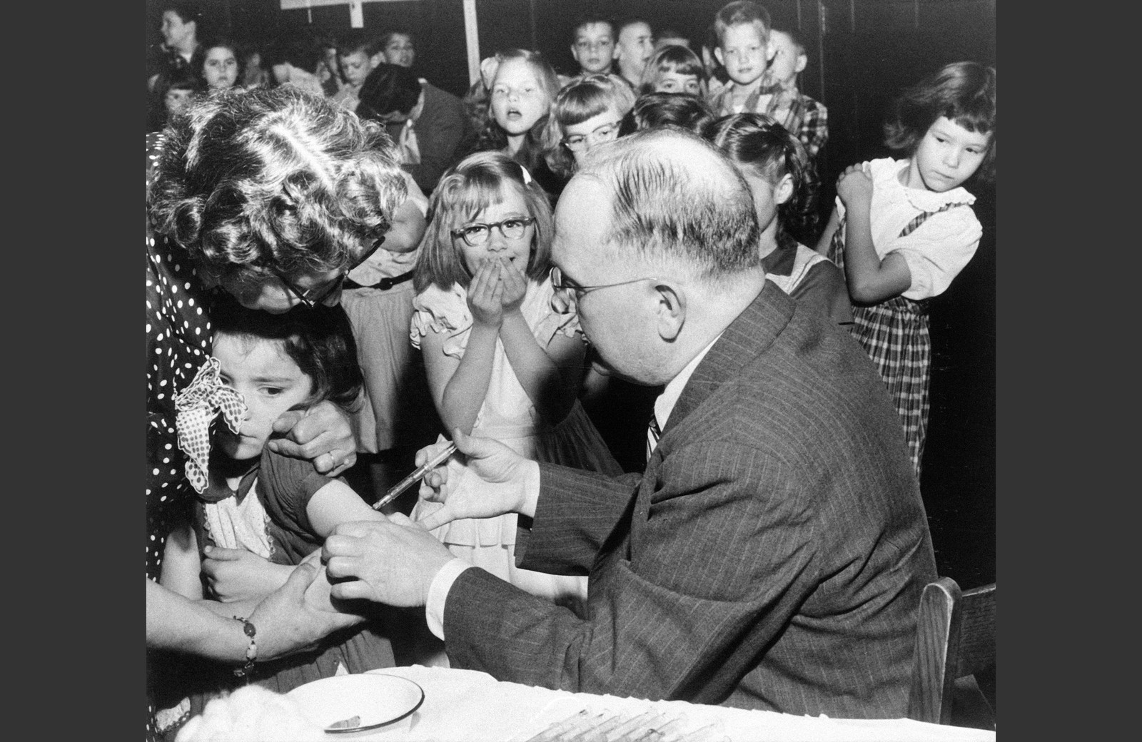 A small crowd of children look on as a girl is given a vaccine shot.