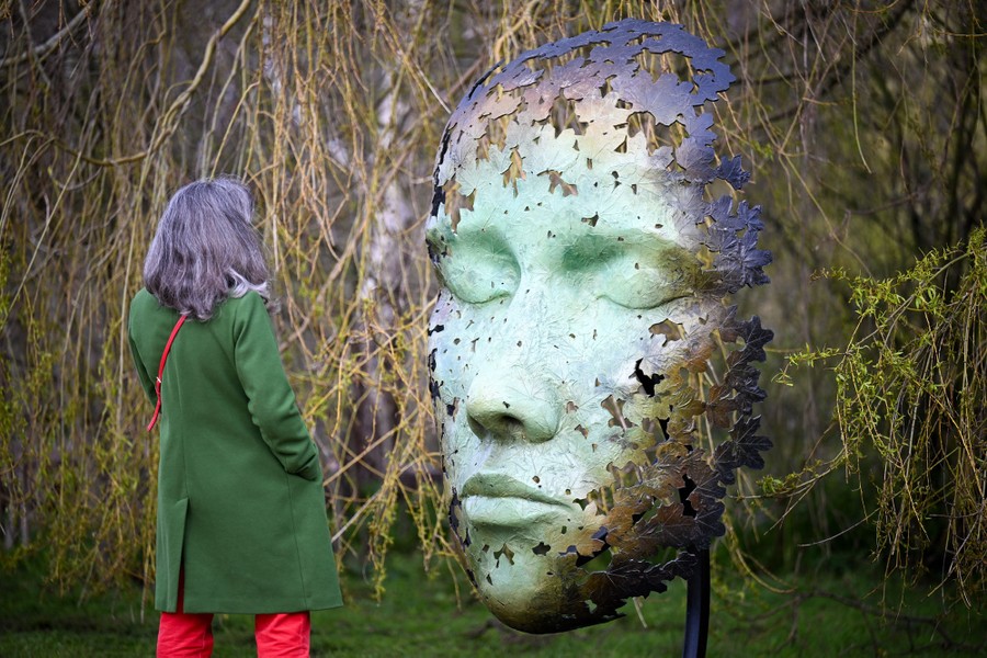 A person stands in front of a sculpture—a large human face—in a park.