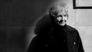 A black-and-white portrait of Alice Munro, standing against a wall and gazing to the right
