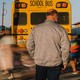A man wearing a medical mask looks into the distance as masked students walk to school buses after class.