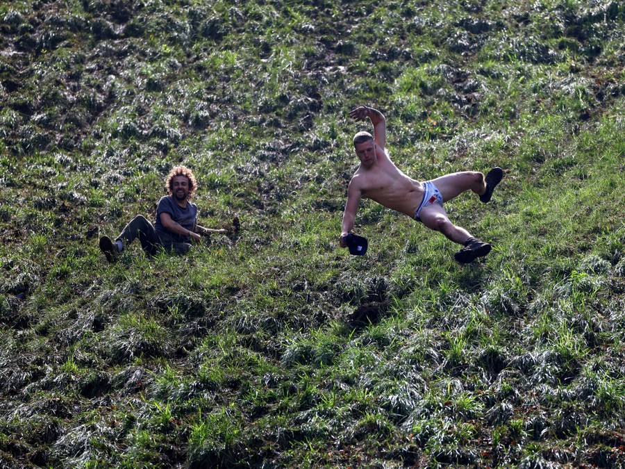 Two men, one of whom wears only briefs and shoes, tumble and slide down a hill.