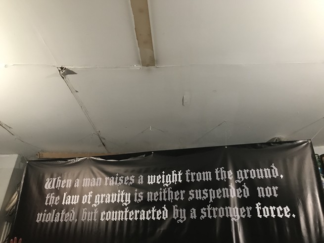 A large sign hanging on the wall of a garage: "When a man raises a weight from the ground, the law of gravity is neither suspended nor violated, but counteracted by a stronger force."
