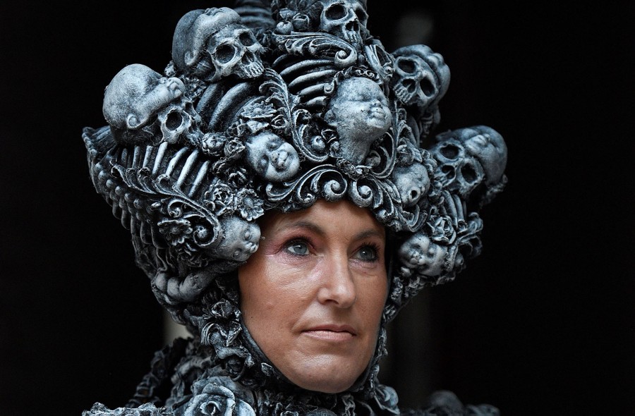A person poses while wearing a head covering made up of small skeletal figures and faces.