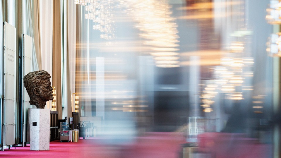 A blurred color photo of the Grand Foyer of the Kennedy Center, showing a bronze bust of John F. Kennedy in focus.