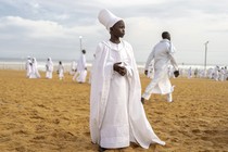 A young person, dressed in a white robe, stands on a beach, with many others dressed in white in the background.