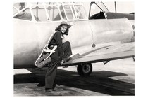 black-and-white archival photo of woman in flight gear and cap, smiling at camera, standing with one foot on ground and one foot on the wing of the plane she piloted, about to climb into cockpit