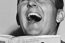 A black-and-white photo of a man throwing his head back in laughter while holding a book