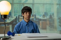 Julio Torres in a blue button down sitting at a table with a giant light bulb lamp