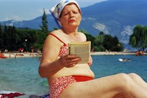 A woman in a kerchief and red bikini reads a book on a beach.