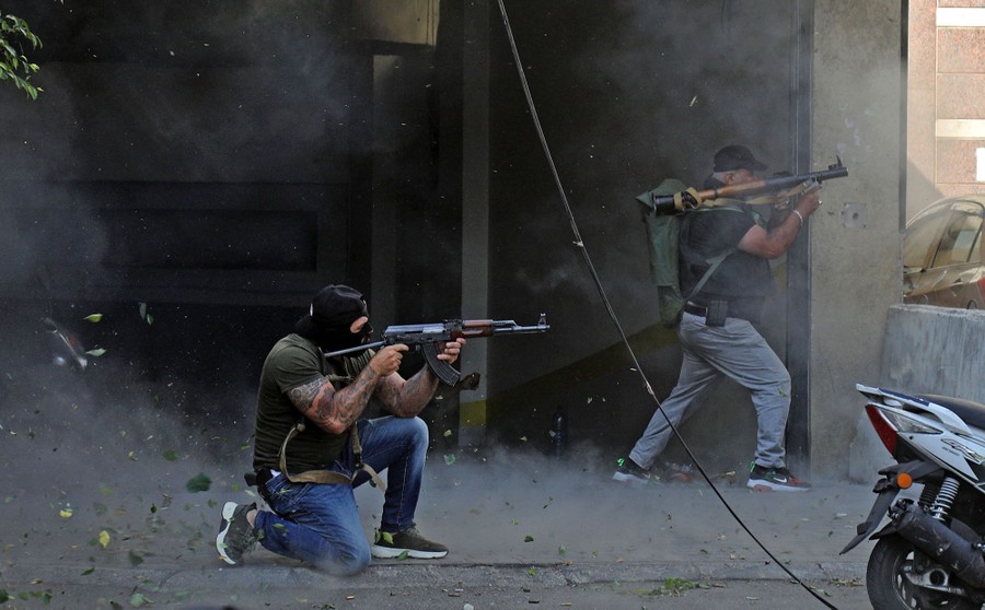 Two men aim weapons down a city street; one kneels and another shelters beside a building. Dust and small bits of debris are flying around.