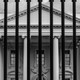 A black and white photograph of the White House through the bars of its fence.