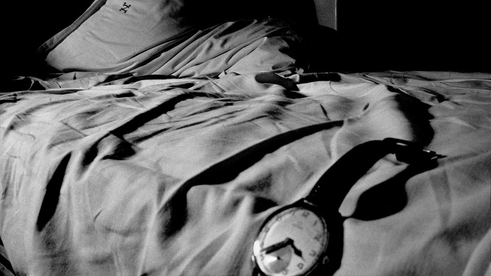 In a black-and-white photo, a watch hangs off an unmade bed cast in shadows.