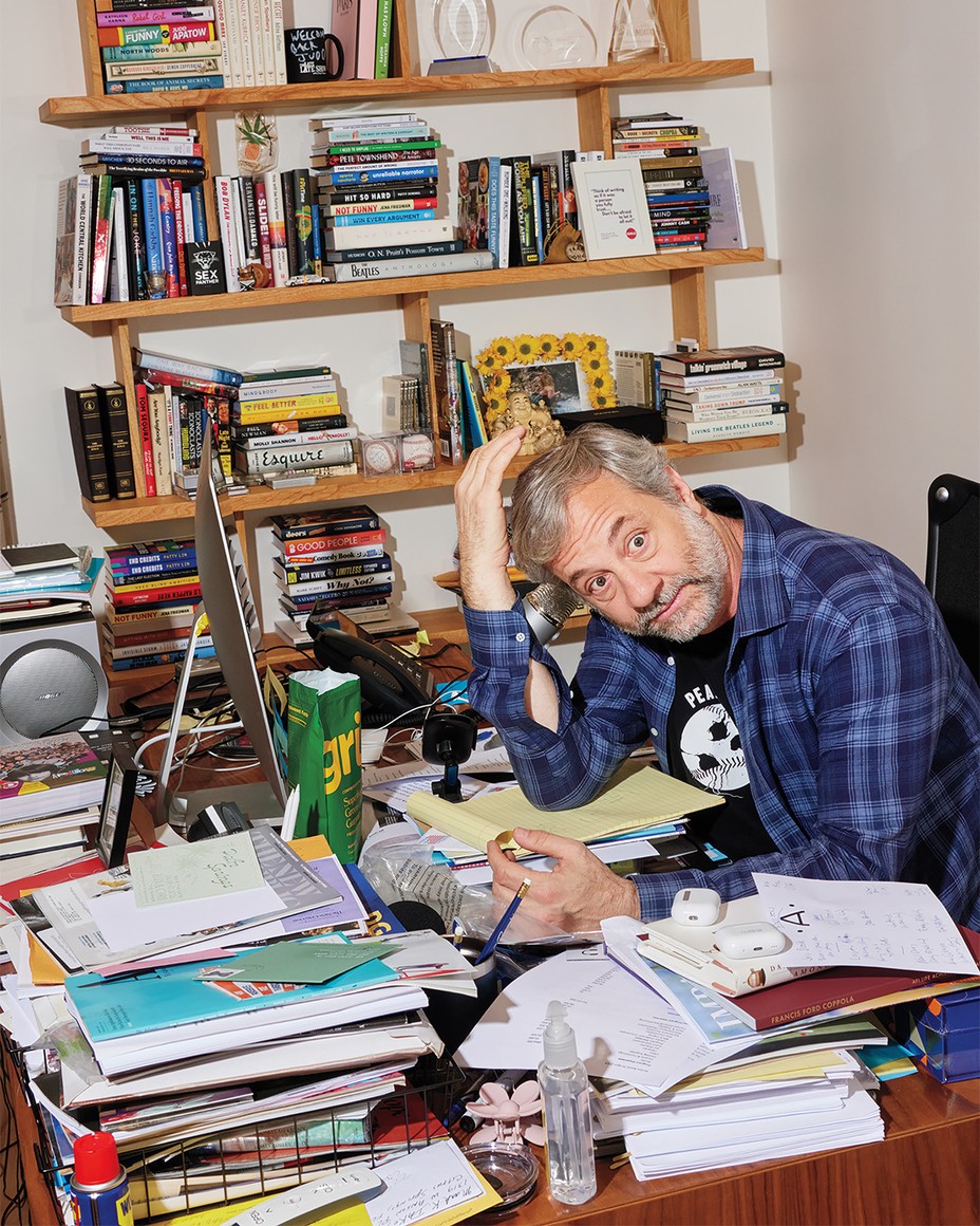 color photo of Apatow at cluttered desk with head in one hand, with bookshelves behind
