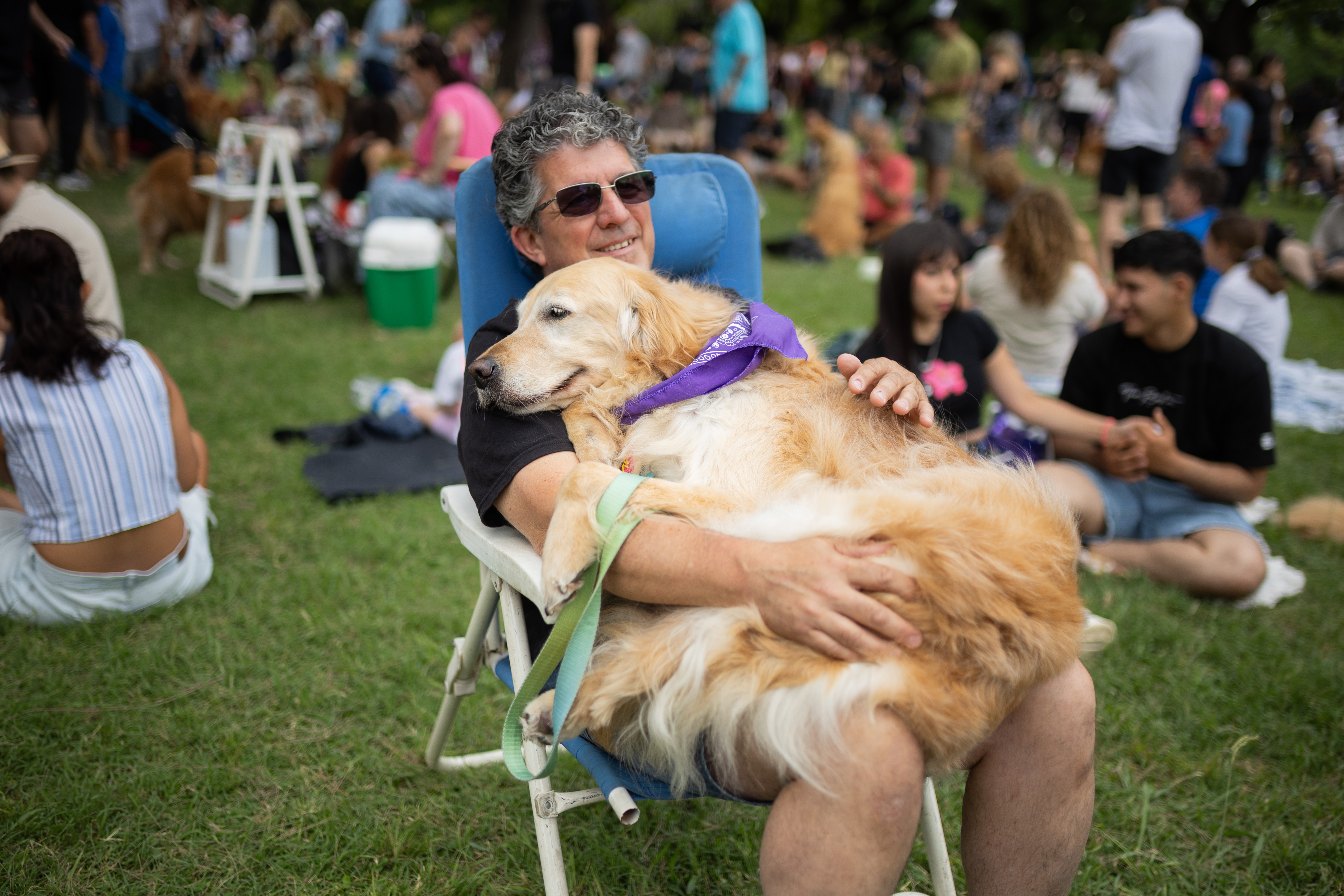 A golden retriever owner sits with his dog on his lap in a park, surrounded by many other people.