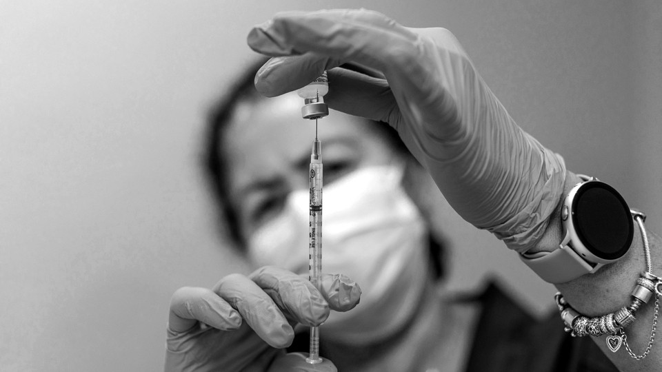 A black and white photo of a woman wearing a mask pulling the COVID-19 vaccine into a syringe with gloved hands