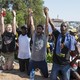 Five young adults sit on their knees, holding hands with their arms above their heads. Their mouths are open as they appear to be yelling.