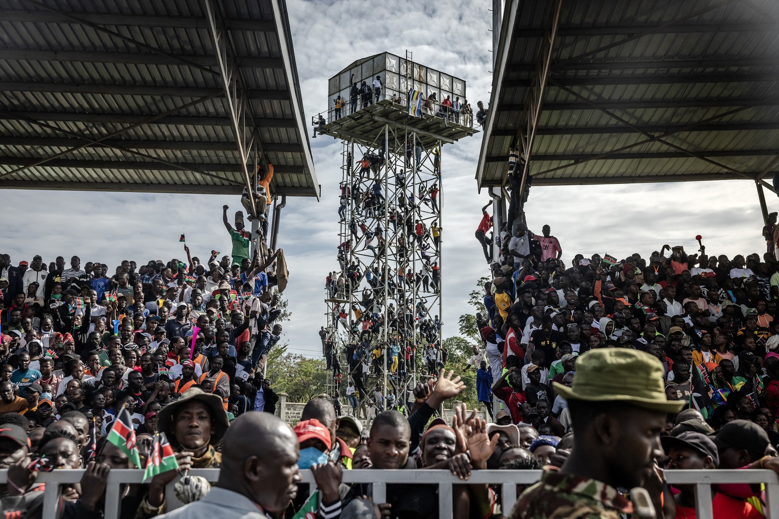 A large crowd fills viewing stands, while many others climb up on a tower nearby.