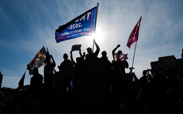 Trump voters raise their fists and wave pro-Trump flags in a protest. A blue sky is seen behind them.