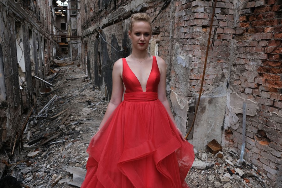 A young woman wears a red gown while standing in a rubble-strewn hallway of her destroyed school building.