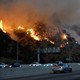A fire burns on a hillside next to a freeway as cars drive by.