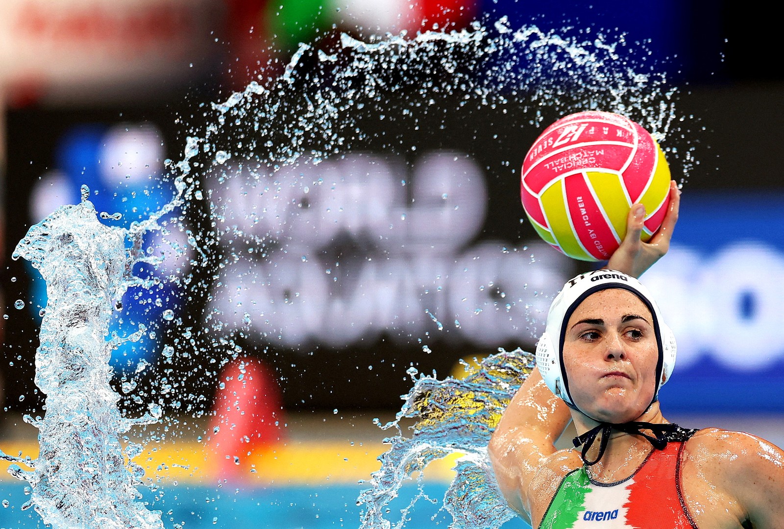 A water polo player raises the ball to make a pass, splashing water in the air.