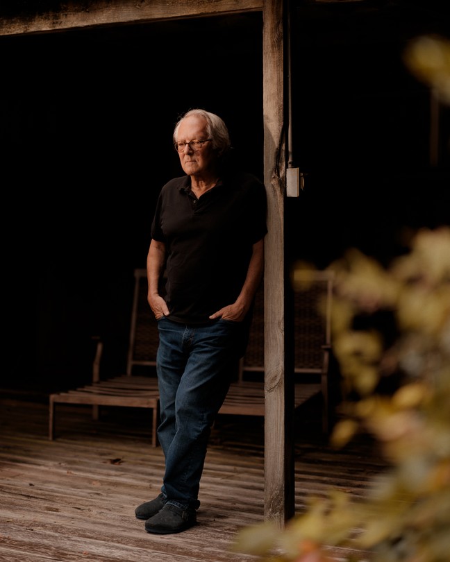 A man wearing a black shirt and jeans leans against a wood post.