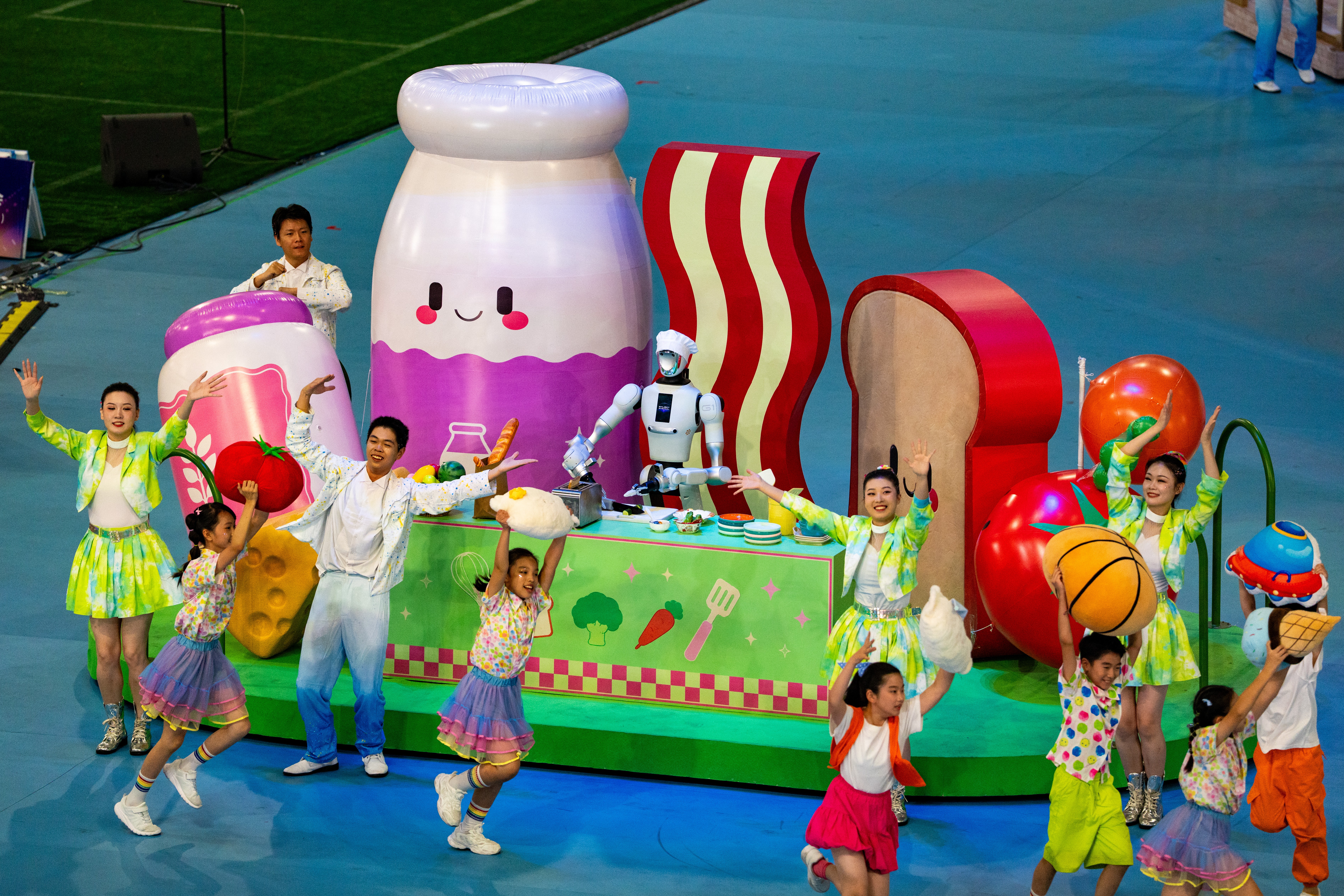 Several human performers dance beside a platform featuring anthropomorphized food, centered around a humanoid robot acting as a chef.