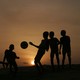 The silhouettes of a group of children kicking a soccer ball in front of the setting sun