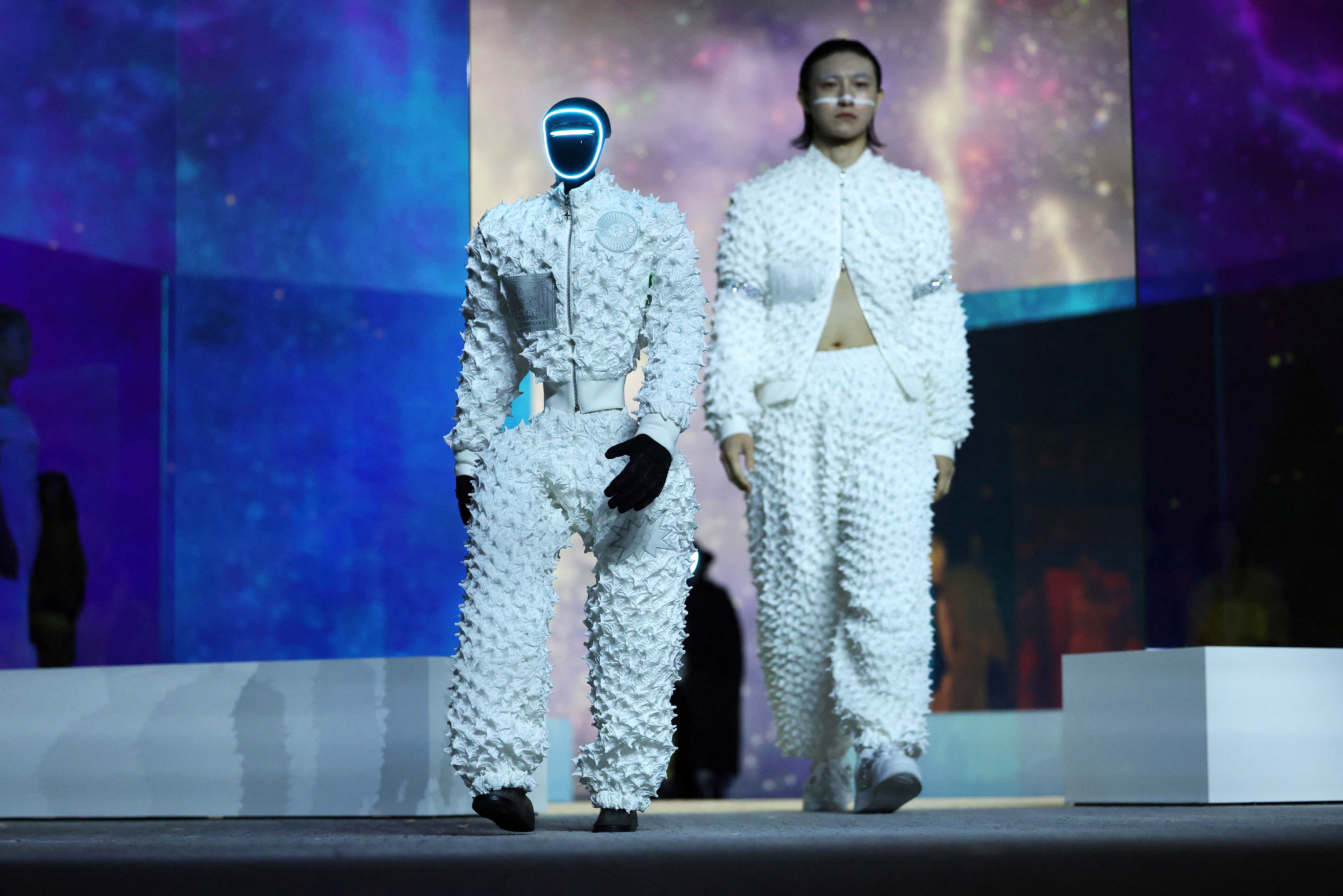 A humanoid robot and a human model, wearing matching garments, walk in a fashion show.