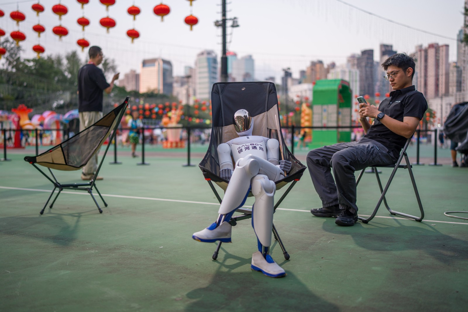 A robot sits, cross-legged, in a chair in a park among humans.