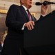 A photograph of Donald Trump speaking into a microphone, with Joe Biden in the background