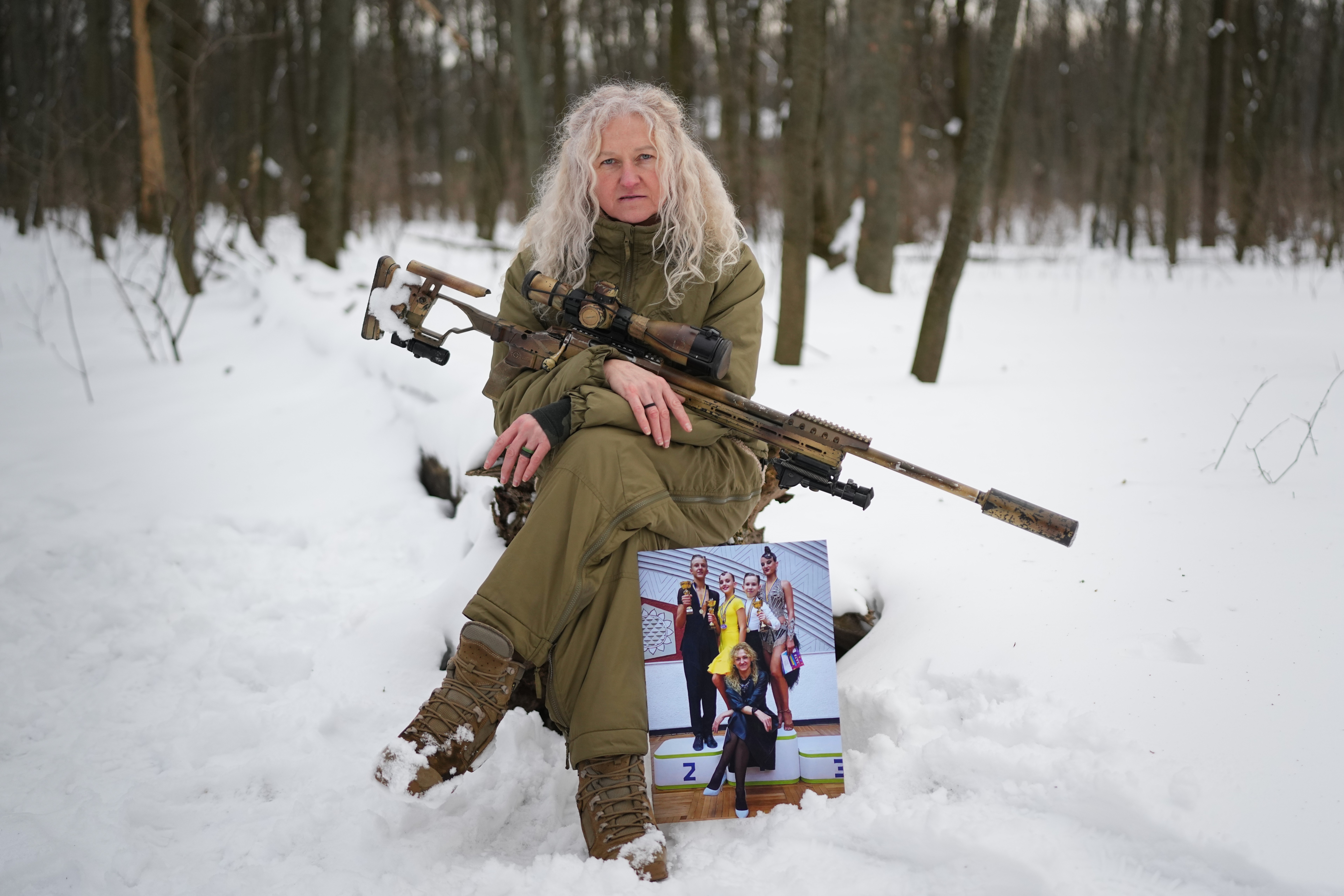 A woman sits on a log in a snowy forest, posing with a sniper rifle across her lap, and a photograph of herself as a dance teacher at her feet.